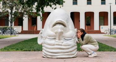 woman next to a sculpture of an upside down head, whispering it its ear