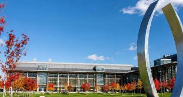 University of California, Merced campus buildings and a large metal arch sculpture framed by autumn trees under a bright blue sky.