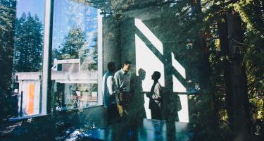 Students gathered inside a glass-walled University of California, Santa Cruz building surrounded by redwood trees.