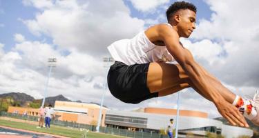 A track athlete stretches toward his shoes on an outdoor field.