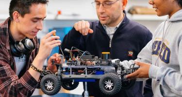 Three people examine a wheeled robotics project on a workbench.