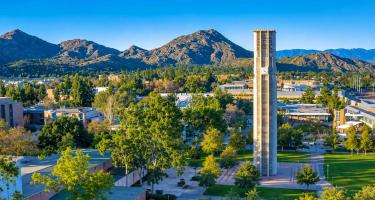 Aerial view of the University of California, Riverside campus with its clock tower, green spaces, athletic fields, and mountains in the background.