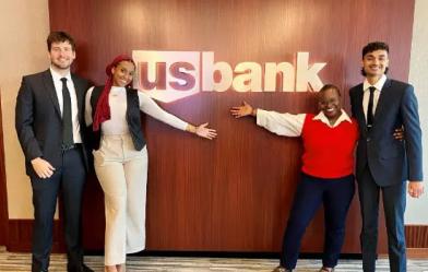 Two interns and two staff members embracing in front of the UC Bank logo on a wall