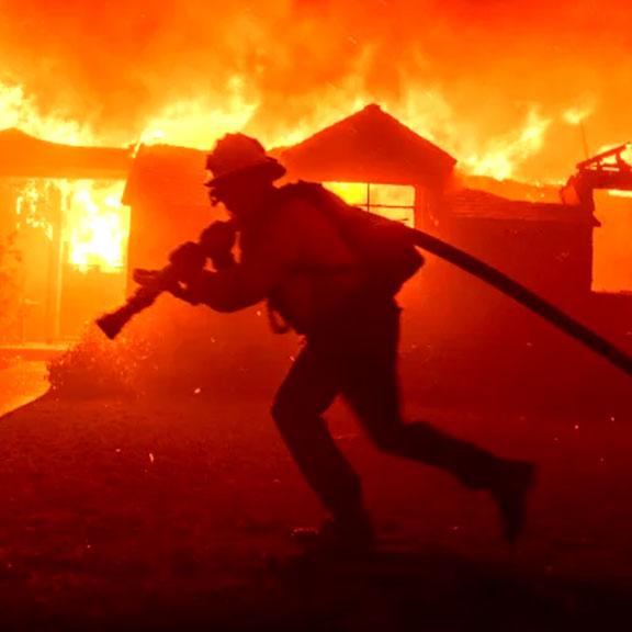 a fireman running in front of a burning house, carrying a firehose
