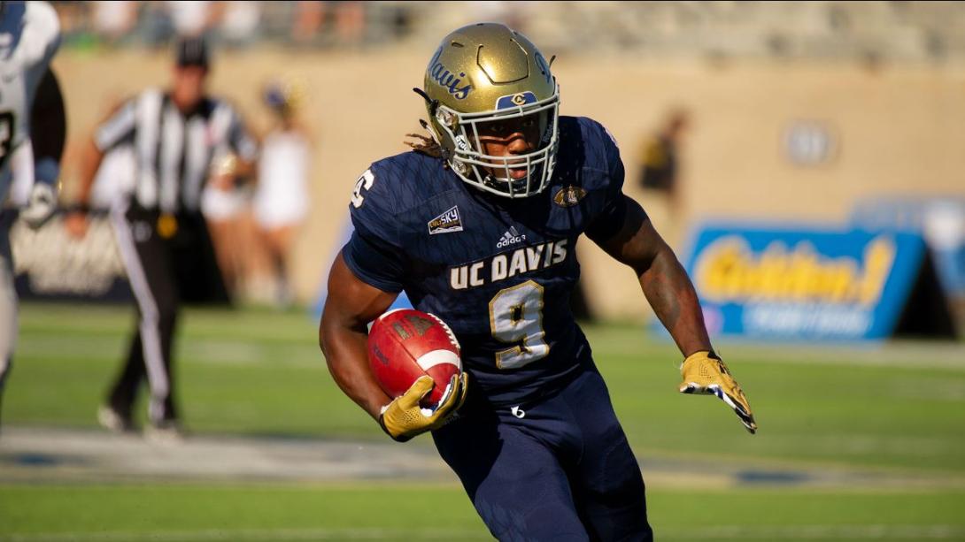 A UC Davis football player holding a football, running across a field