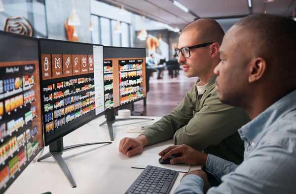 Two men sitting in front of three monitors, discussing strategy